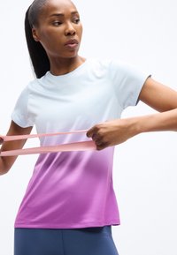 Γυναίκα που φοράει μπλουζάκι γυμναστικής με βαθμιαία απόχρωση του μωβ τεντώνει μια ροζ ελαστική ταινία, δείχνοντας συγκεντρωμένη μπροστά σε απλό λευκό φόντο.