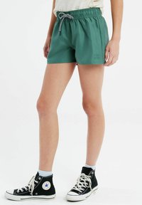 Shorts verts, légers, avec une taille élastique et un cordon de serrage, arborant un petit logo de marque sur le côté. Associés à des baskets montantes noires.