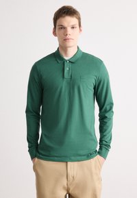 Polo cu mânecă lungă, de culoare verde, fabricat dintr-un material moale, având guler, placket cu trei butoane și o mică etichetă de marcă pe piept.
