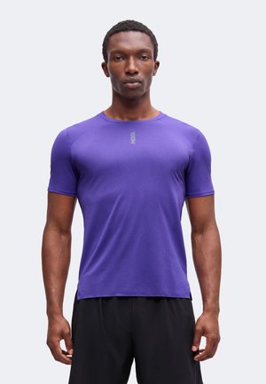 Homme aux cheveux courts et bouclés portant une chemise violette ajustée à manches courtes et un pantalon noir, debout devant un fond clair uni.