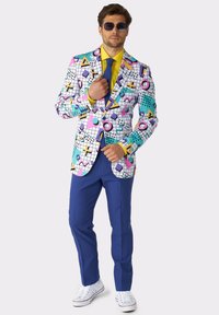 Giacca da uomo colorata con motivi geometrici in rosa, blu e giallo, abbinata a pantaloni blu e sneaker bianche.