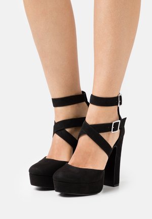 Chaussures à talons hauts en suède noir avec une plateforme, dotées de brides croisées autour de la cheville et de boucles carrées, présentant une texture lisse et un design élégant.