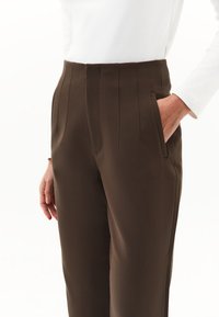 Donna che indossa pantaloni a vita alta marroni, sagomati, con cuciture verticali, mano destra nella tasca, abbinati a un top bianco a maniche lunghe.