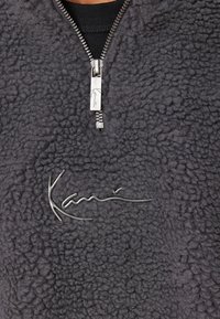 Mørkegrå fleece-pullover med halvlukning med lynlås, tekstureret overflade og en metallisk logo på fronten.