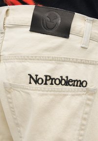 Κοντινή λήψη της πίσω τσέπης σε ανοιχτό μπεζ παντελόνι με μαύρο κεντητό κείμενο "No Problemo" και μαύρο δερμάτινο αυτοκόλλητο πάνω από την τσέπη.