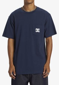 Tmavě modré bavlněné tričko s krátkým rukávem, kapsou a bílým potiskem loga. Mírně volnější střih, kulatý výstřih, hladká textura.