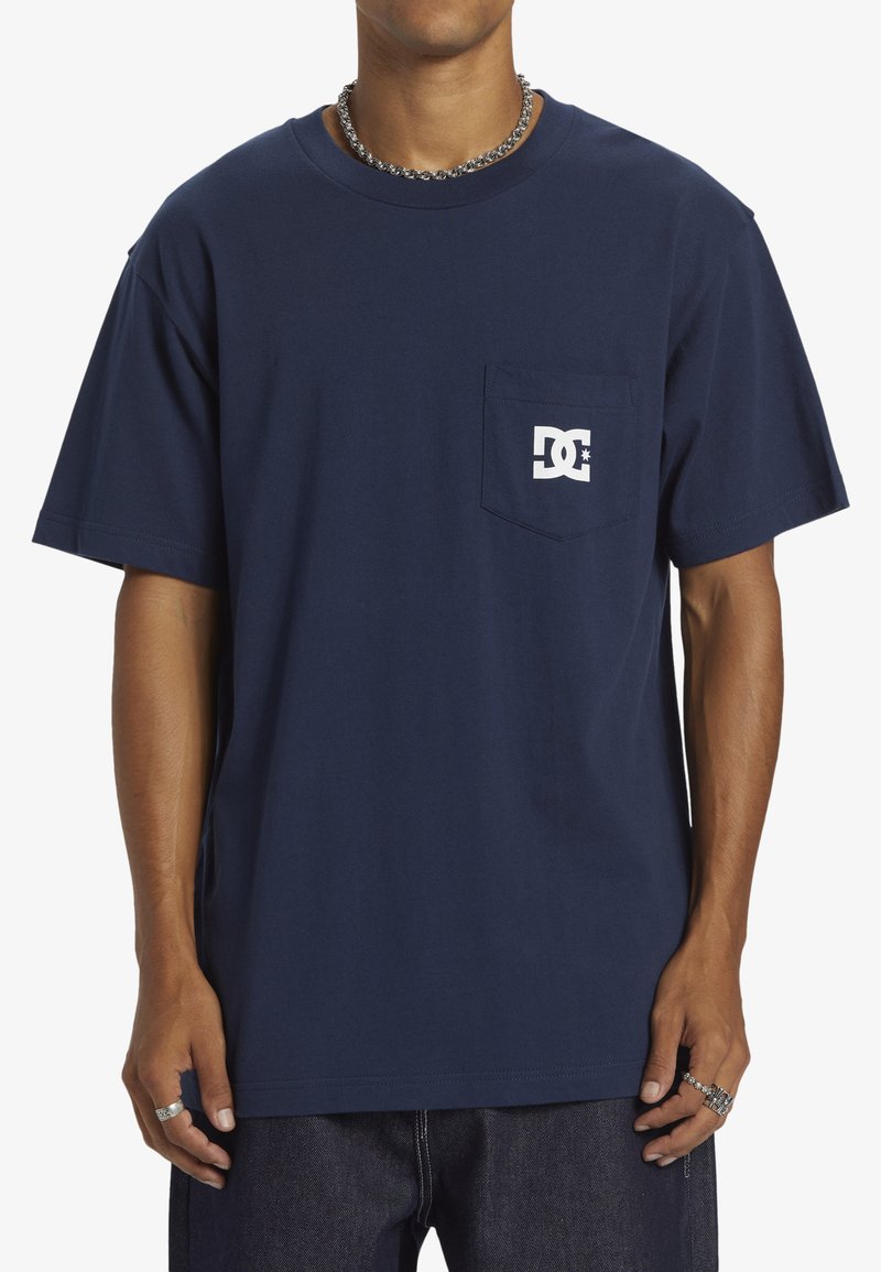 Tmavě modré bavlněné tričko s krátkým rukávem, kapsou a bílým potiskem loga. Mírně volnější střih, kulatý výstřih, hladká textura.