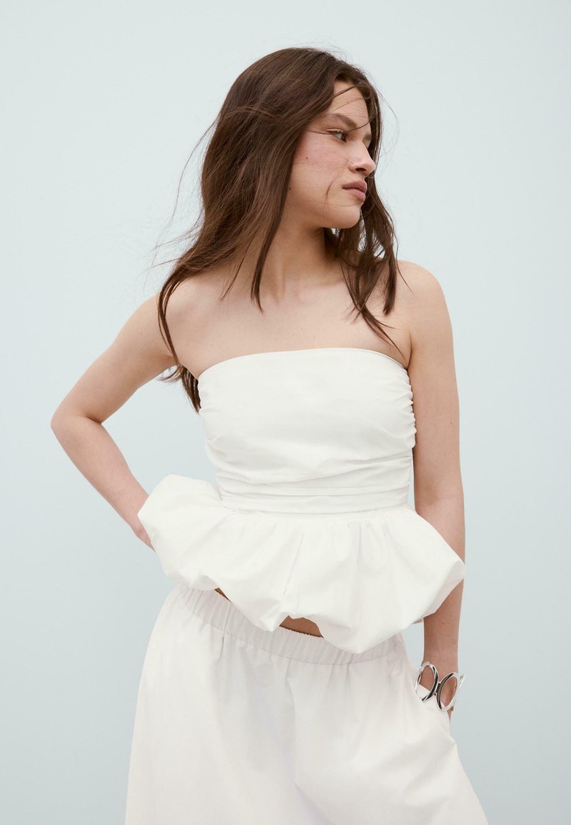 Femme aux longs cheveux bruns portant un haut peplum blanc sans bretelles et un pantalon blanc, posant la main sur la hanche sur un fond clair.