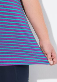 Tunika pruhovaná v purpurové a tyrkysové barvě s uvolněným střihem, vyrobená z měkké bavlny a s zakřiveným spodním lemem, částečně držená v ruce.