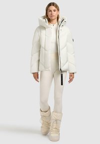 Witte pufferjas met een hoge kraag, ruitvormige quiltontwerp en ritssluiting. G gedragen over bijpassende witte leggings en lichtgekleurde laarzen.