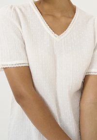 Blusa blanca de manga corta con cuello en V, tela texturizada y ribete de encaje decorativo en el escote y las mangas, con pequeños estampados de puntos.