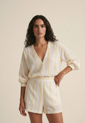 Mujer con cabello largo y rizado que lleva una blusa de lino blanca y pantalones cortos de talle alto a juego con rayas verticales amarillas, de pie con una mano en la cadera.