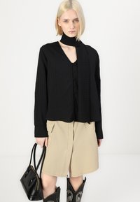 Camicia nera con bottoni, a righe verticali, maniche lunghe e colletto con sciarpa annodata, abbinata a una gonna beige fino al ginocchio e a una borsa nera.