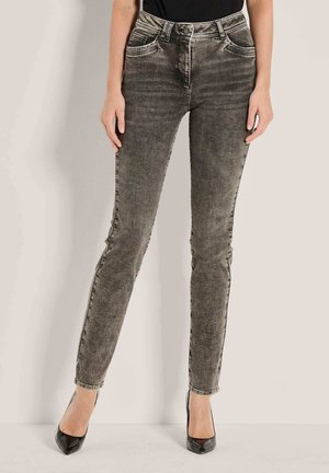 Vrouw in strakke vervaagde zwarte jeans en zwarte puntige hakken, staand tegen een effen lichte achtergrond.