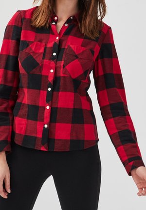 Kobieta ubrana w czerwoną i czarną koszulę w kratę na guziki z dwoma kieszeniami na piersi, w połączeniu z czarnymi spodniami, na jednolitym tle.