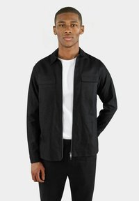 Svart zip-up jacka i ett mjukt tyg med två bröstfickor och en klassisk krage, bärs över en vit t-shirt, tillsammans med svarta byxor.