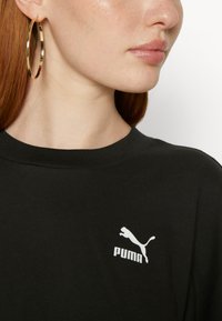 Black cotton t-shirt with a crew neck and short sleeves, featuring a white Puma logo on the left chest area. Model has gold hoop earrings.