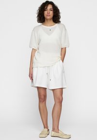 Top corto a maniche corte in maglia bianca e testurizzata abbinato a pantaloni corti bianchi con cordoncino. Le scarpe slip-on di colore crema completano l'outfit.