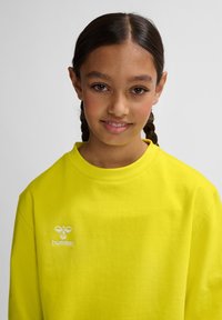 Klart gul sweatshirt gjort av ett mjukt material. Har en rund hals och en liten vit "hummel"-logotyp broderad på framsidan.