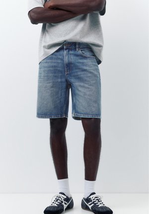 Mann trägt hellblaue Jeans-Shorts, graues T-Shirt, weiße Socken und schwarz-weiße Sneakers und steht mit verschränkten Armen.