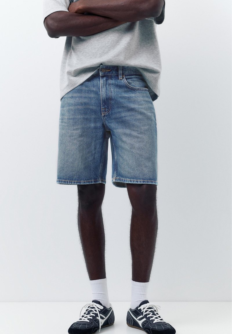 Homme portant un short en jean bleu clair, un t-shirt gris, des chaussettes blanches et des baskets noires et blanches, debout les bras croisés.