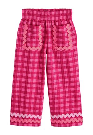 Onderbenen met roze geruite broek met golvende zoom en witte sandalen met gekruiste bandjes op een lichte vloerachtergrond.
