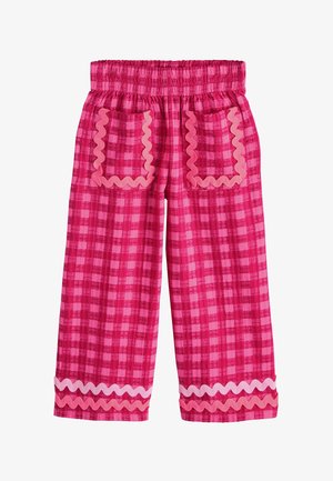 Unterschenkel mit rosa karierter Hose mit welligem Saum und weißen Sandalen mit gekreuzten Riemen auf hellem Boden.