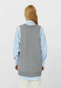 Femme aux longs cheveux blonds portant une chemise bleu clair sous un gilet en tricot gris sans manches, se tenant de dos devant un fond uni.