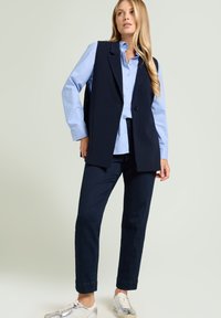 Marineblauwe mouwloze blazer over een lichtblauw gestreept overhemd, gecombineerd met donker denimbroek en zilveren sneakers. Gladde stof met een getailleerde pasvorm.