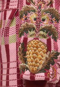 Κόκκινο και ροζ καρό ύφασμα που διαθέτει λεπτομερείς κεντητές σχεδιάσεις ανανά και λουλουδιών με πράσινες και χρυσές λεπτομέρειες σε υφασμάτινη επιφάνεια.