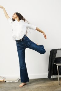 Bela dolga majica s long sleeves in modri široki kavbojki z črnim pasom. Manekenka se dinamično poda bosa proti nevtralni ozadju.