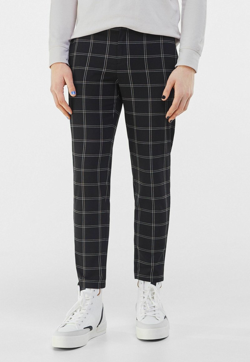 Černé kárované kalhoty s bílými a světle šedými proužky, úzkého střihu, s hladkou texturou. Kombinovány s bílými vysokými teniskami.