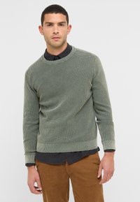 Sweatshirt en velours côtelé vert avec un col rond, surface texturée et manches longues, superposé à une chemise à col sombre.