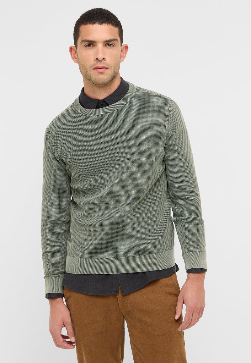 Sweatshirt en velours côtelé vert avec un col rond, surface texturée et manches longues, superposé à une chemise à col sombre.