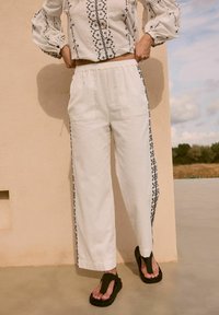 Pantaloni larghi in lino color avorio con dettagli ricamati neri sui lati. Abbinati a infradito scure. Dotati di una cintura elasticizzata.