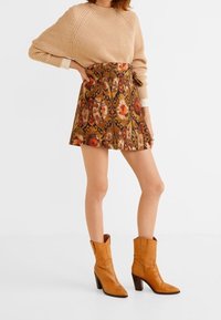 Beige neulepaita, kukkakuosiset ruskeat hameet ja vaaleat cowboy-kengät, joissa on pinottu korko. Asu sisältää teksturoituja materiaaleja ja eloisia värejä.