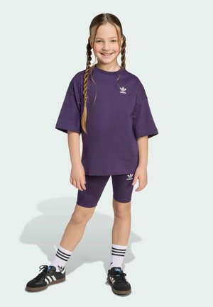 Lila Baumwoll-T-Shirt und passende Fahrradshorts mit weißem Adidas-Logo; kombiniert mit schwarzen Turnschuhen und weißen gestreiften Socken.