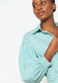 Camicia a strisce verdi e bianche con colletto classico, maniche a sbuffo e polsini abbottonati. Tessuto liscio con vestibilità strutturata.