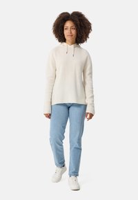 Helle beigefarbene Strick-Hoodie mit einer lockeren Passform, Kordelzugdetails und langen Ärmeln, kombiniert mit hellblauen Jeans und weißen Sneakers.