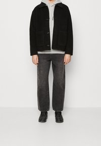 Musta villasekoitekankainen takki napin sulkemisella ja kahdella sivutaskulla, kerroksittain harmaan hupparin päällä, yhdistettynä tummanharmaisiin farkkuihin ja mustiin lenkkareihin.