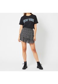 Černé grafické tričko s potiskem "NEW YORK", spárované s černou květinovou mini sukni s volánkovým lemem. Modelka má na sobě černé šněrovací boty.