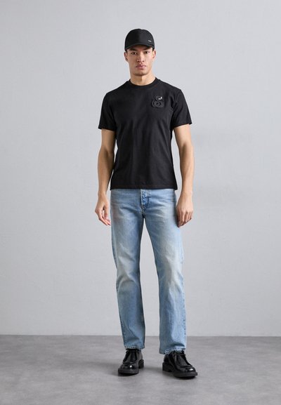 Zwarte katoenen t-shirt met korte mouwen en een logo op de borst, gecombineerd met lichtblauwe denim jeans en zwarte veterschoenen.