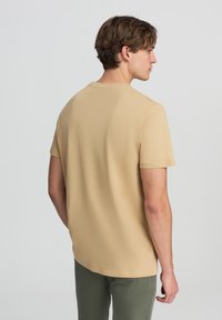 T-shirt a maniche corte, di un leggero color beige, realizzata in tessuto morbido, con scollatura rotonda e orlo dritto, indossata con pantaloni verdi.