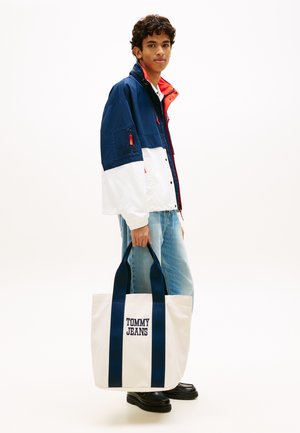 Joven con chaqueta azul marino, blanca y roja y vaqueros, sosteniendo una bolsa de tela blanca con asas azul marino y el logo "Tommy Jeans".