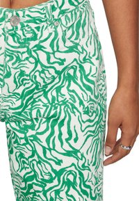 Calças de denim verde e branco com padrões e riscas onduladas, apresentando botão de metal e bolsos frontais, realçando a superfície texturizada do tecido.