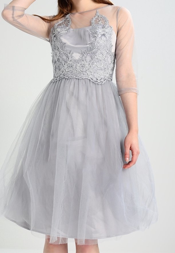 Femme portant une robe gris clair longueur genou avec des manches transparentes et des détails en dentelle complexes sur le corsage.