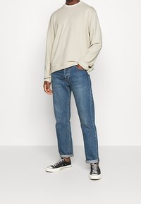 Sudadera gris clara con cuello redondo, combinada con vaqueros de mezclilla azules y zapatillas negras con suelas blancas. Ajuste casual, con un detalle mínimo.