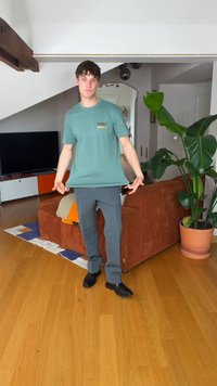 Νεαρός άνδρας φοράει πράσινο T-shirt, τεντώνεται και στέκεται σε ξύλινο πάτωμα σε καθιστικό με πορτοκαλί καναπέ και μεγάλο φυτεμένο φυτό.