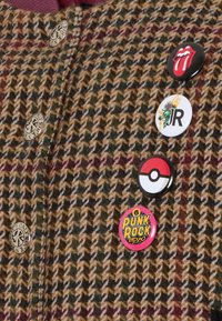 Brązowa tkanina w wzór houndstooth z wielokolorowymi nitkami, ozdobiona pięcioma emaliowanymi przypinkami: Rolling Stones, JR, Pokémon oraz dwoma wzorami punk rock.