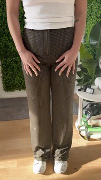 Una persona con pantalones marrones con tachuelas y zapatos blancos está de pie en un interior cerca de plantas verdes y una pequeña mesa con cámaras y libros.